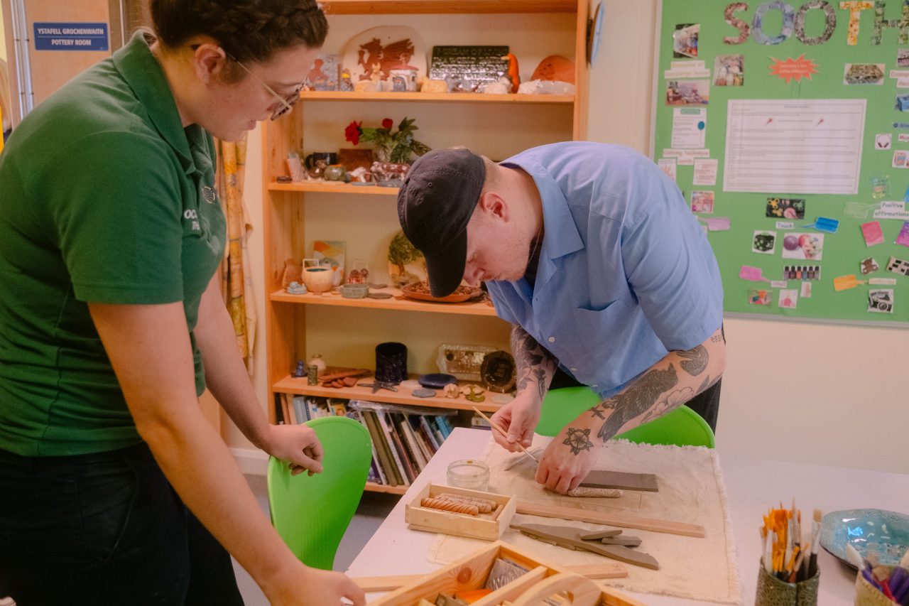 Delwedd o Ystafell Ddillad, aelod staff Bwrdd Iechyd Addysgu Powys yn goruchwylio dosbarth gwneud pottery.