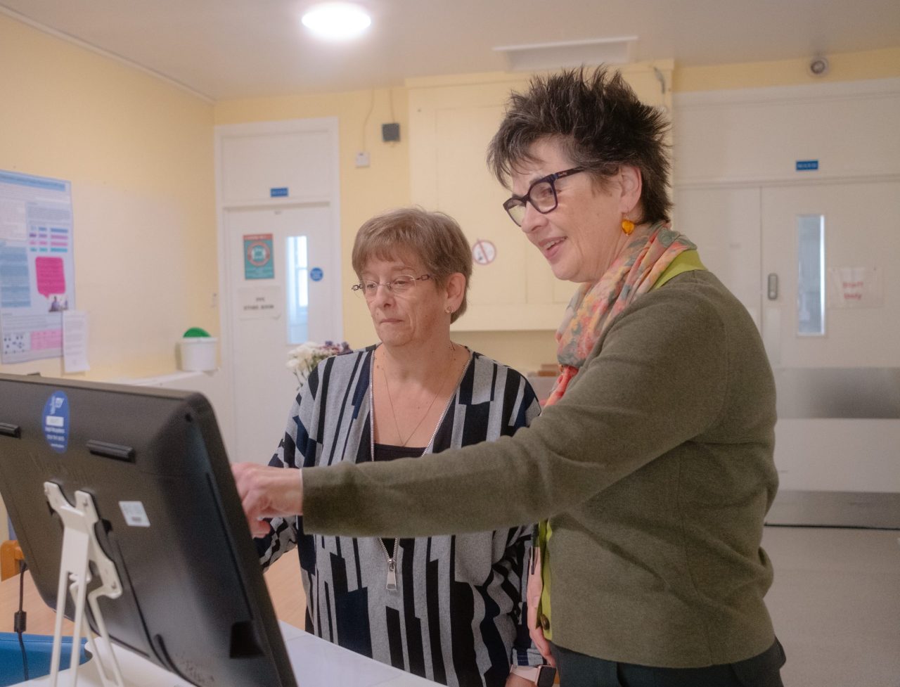 Dau aelod o staff Bwrdd Iechyd Addysgu Powys yn defnyddio cyfrifiadur mewn gosodiad swyddfa