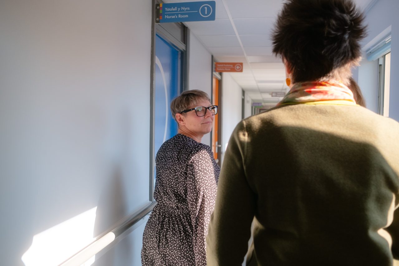 Dau aelod o staff Bwrdd Iechyd Addysgu Powys yn cerdded i lawr coridor, un yn edrych yn ôl ar y llall.