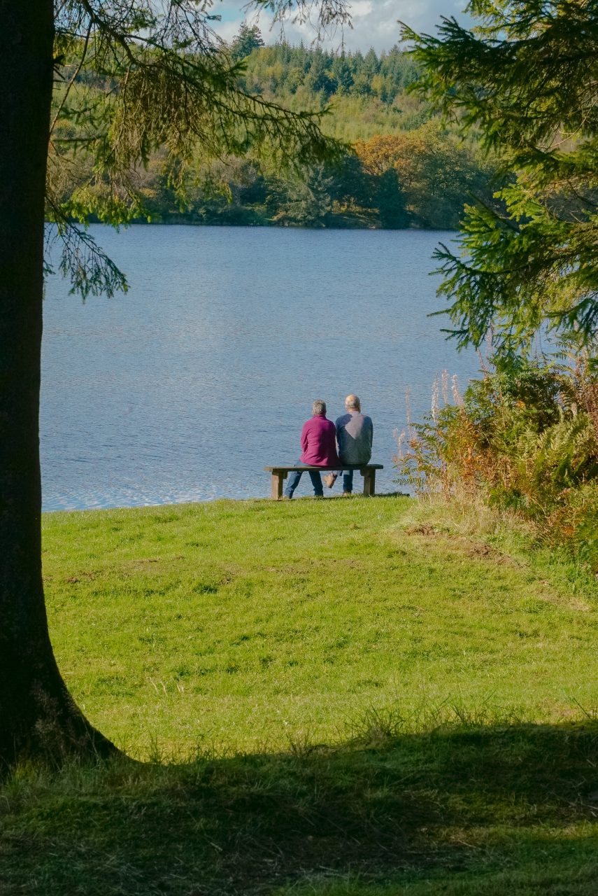 Pâr yn eistedd ar fainc gyda golygfa hardd o llyn Powys.
