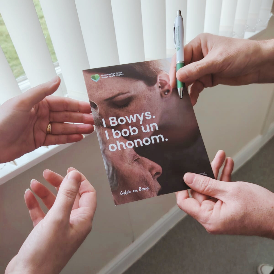 Delwedd o booklet gwybodaeth Elusen Iechyd Powys a phensil yn cael ei rhoi i rywun