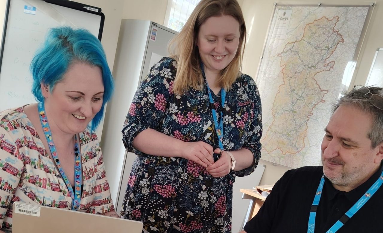 Cyllid agos o Dîm Elusennau Iechyd Powys mewn sefydliad swyddfa gyda llaptopau.